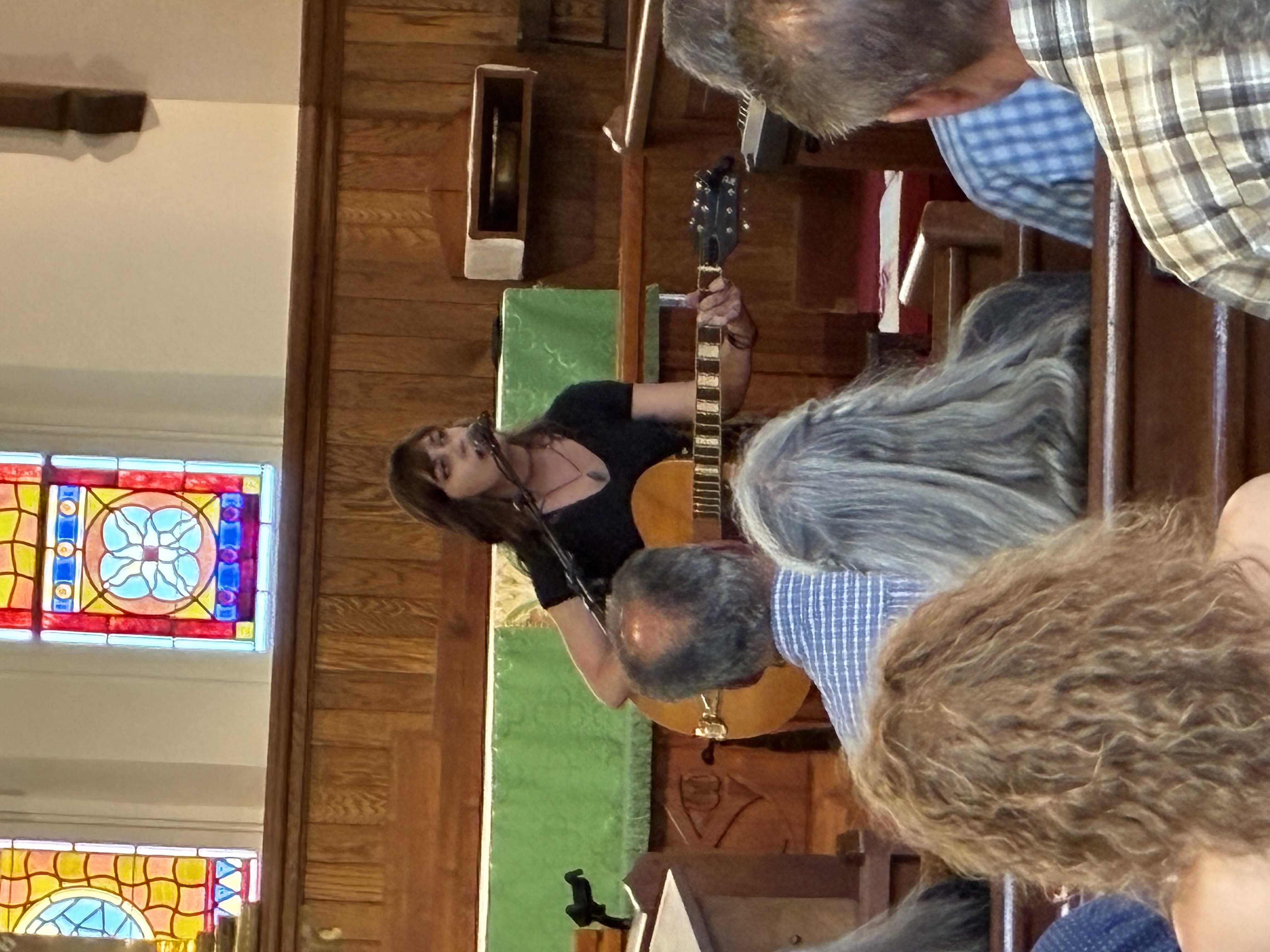 Diana New performing with her guitar at the microphone in front of the stained-glass windows