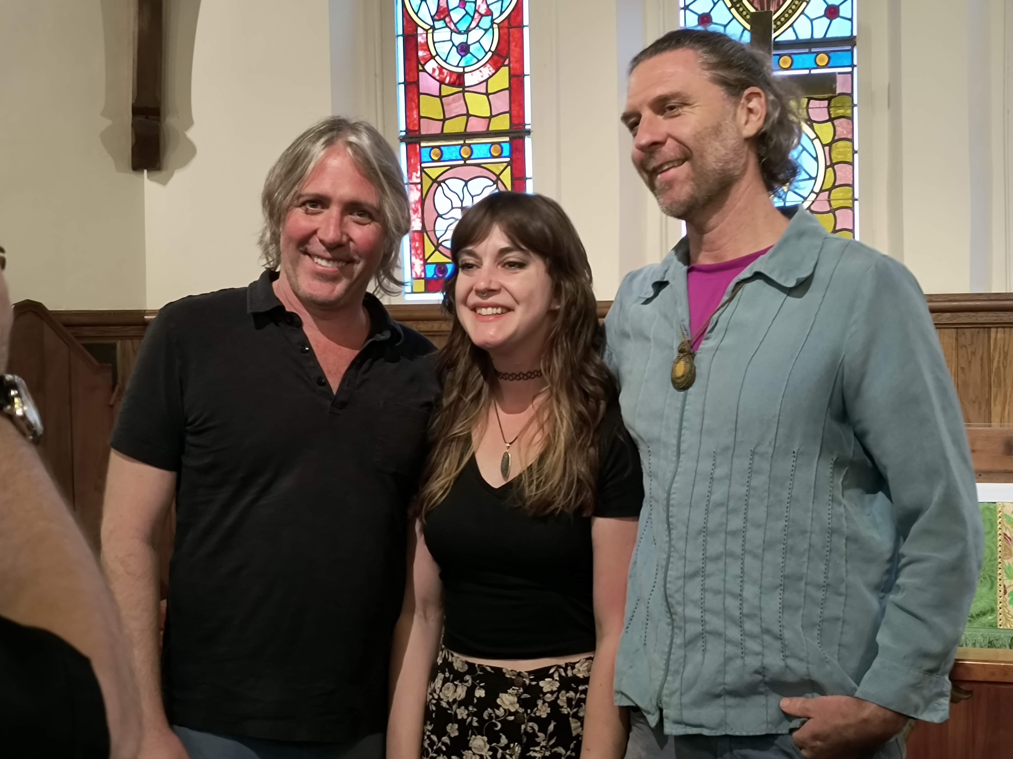 The three featured musicians smiling together in the chapel by the stained-glass windows