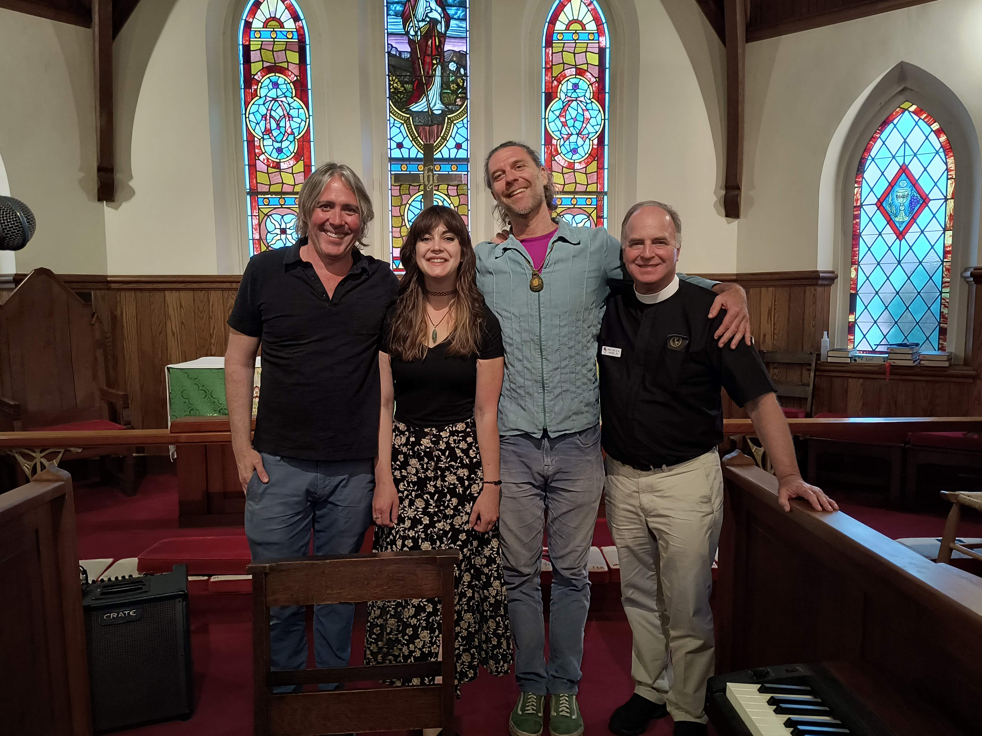 The three performers standing with Fr. in the chapel in front of the stained-glass windows after the concert